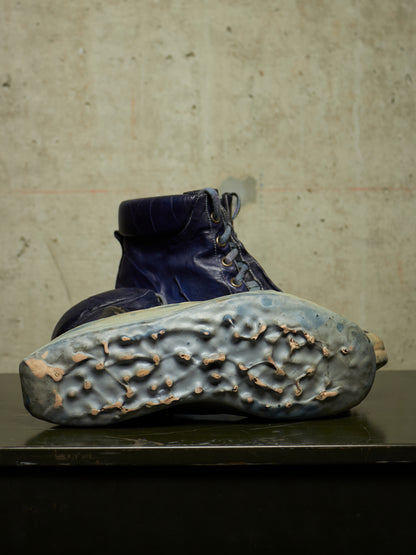 A single dark blue lace-up boot shown from the sole side, resting on a dark surface against a textured concrete wall.