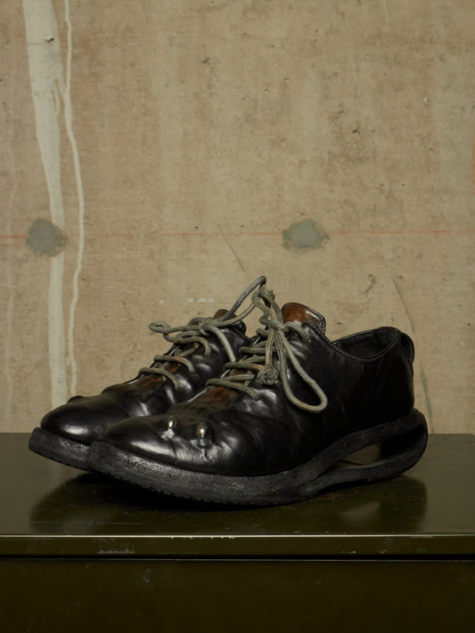 A pair of black leather lace-up shoes with metal pins on the toe boxes and dark soles, displayed on a dark green surface against a textured beige wall.