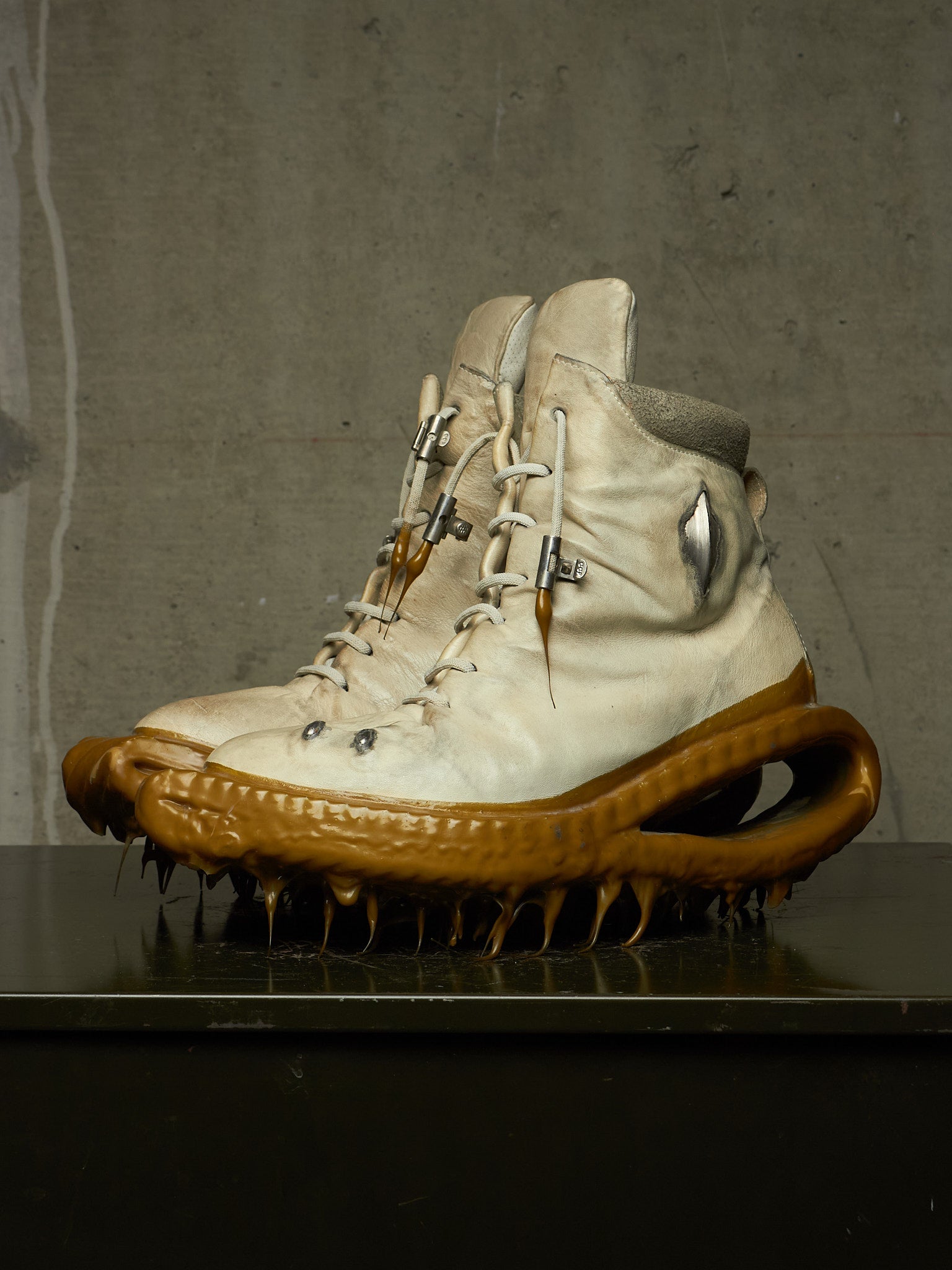 A pair of high-top off-white leather boots with an organic, sculptural brown sole featuring sharp spikes and a looped arch, displayed on a dark reflective surface against a textured gray concrete wall.