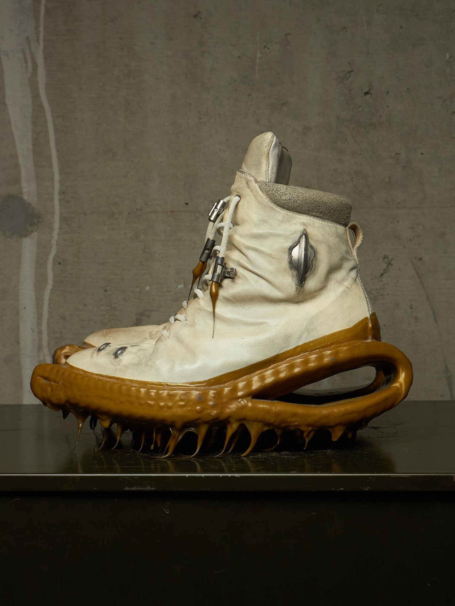 A single high-top sneaker with a cream-colored upper, a thick sculptural brown sole, and metallic hardware, displayed on a dark surface against a textured gray wall.