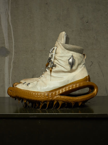 A single high-top sneaker with a cream-colored upper, a thick sculptural brown sole, and metallic hardware, displayed on a dark surface against a textured gray wall.
