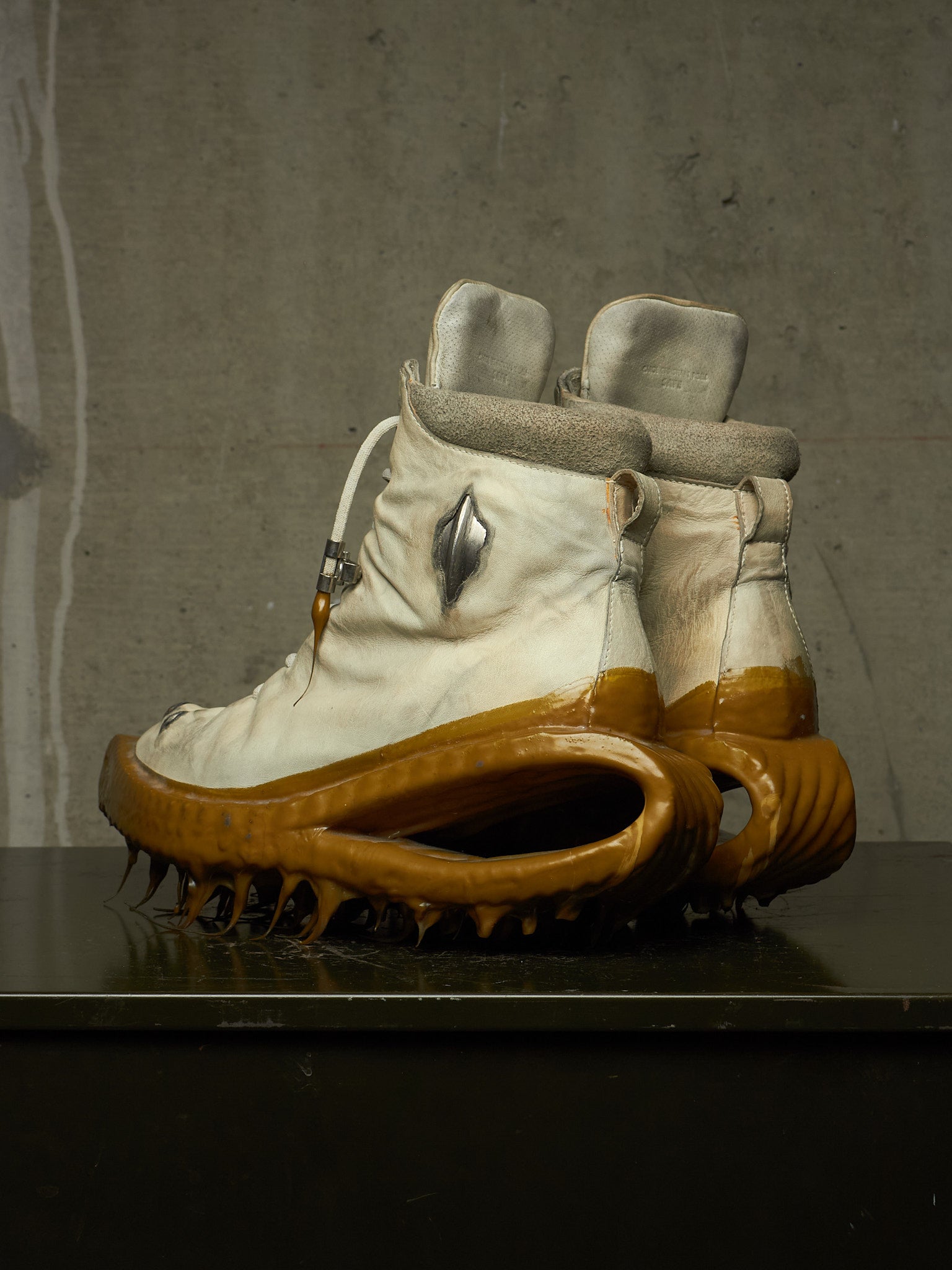 A pair of high-top sneakers with off-white canvas uppers, gray fuzzy trim, and chunky sculptural brown and gold soles, displayed on a dark reflective surface against a textured gray wall.