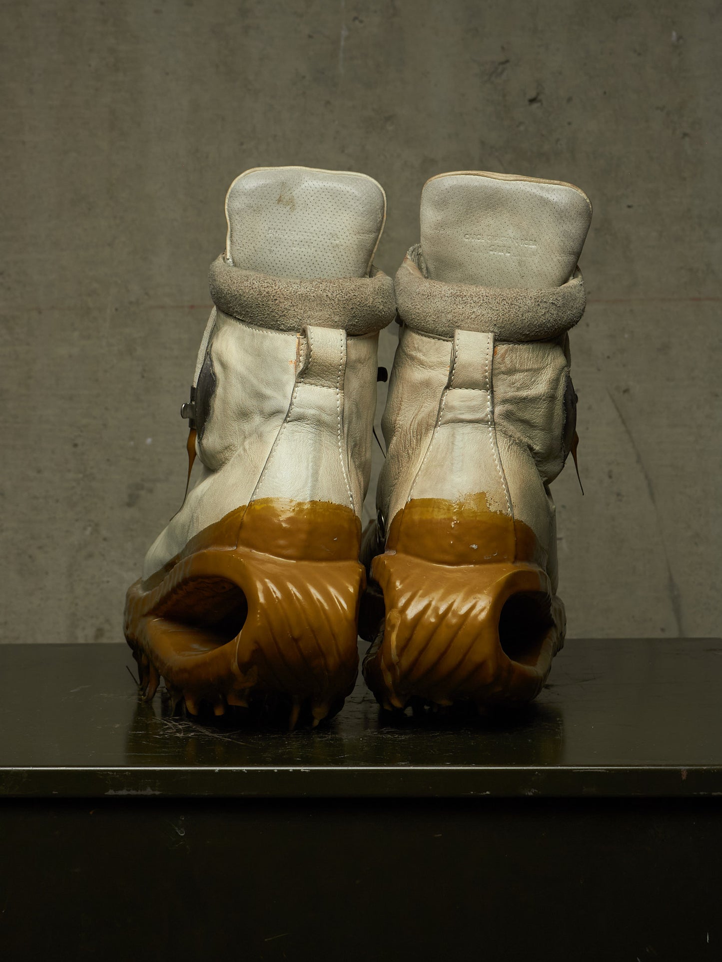 A pair of high-top boots shown from the back, featuring off-white leather uppers with fuzzy trim and chunky brown sculptural soles, resting on a dark surface against a textured gray wall.