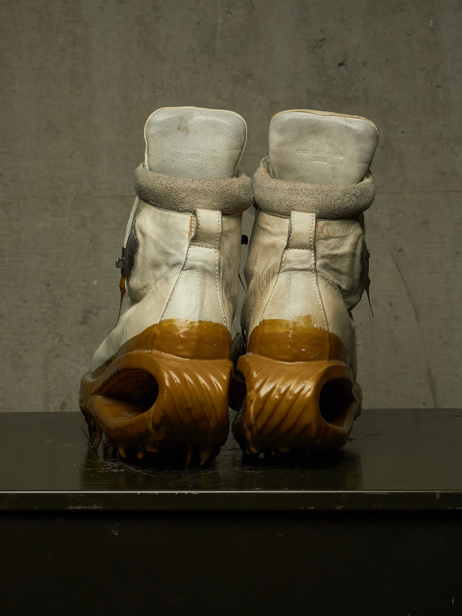 A pair of high-top boots shown from the back, featuring off-white leather uppers with fuzzy trim and chunky brown sculptural soles, resting on a dark surface against a textured gray wall.