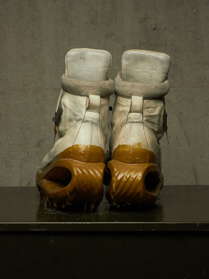 A pair of high-top boots shown from the back, featuring off-white leather uppers with fuzzy trim and chunky brown sculptural soles, resting on a dark surface against a textured gray wall.