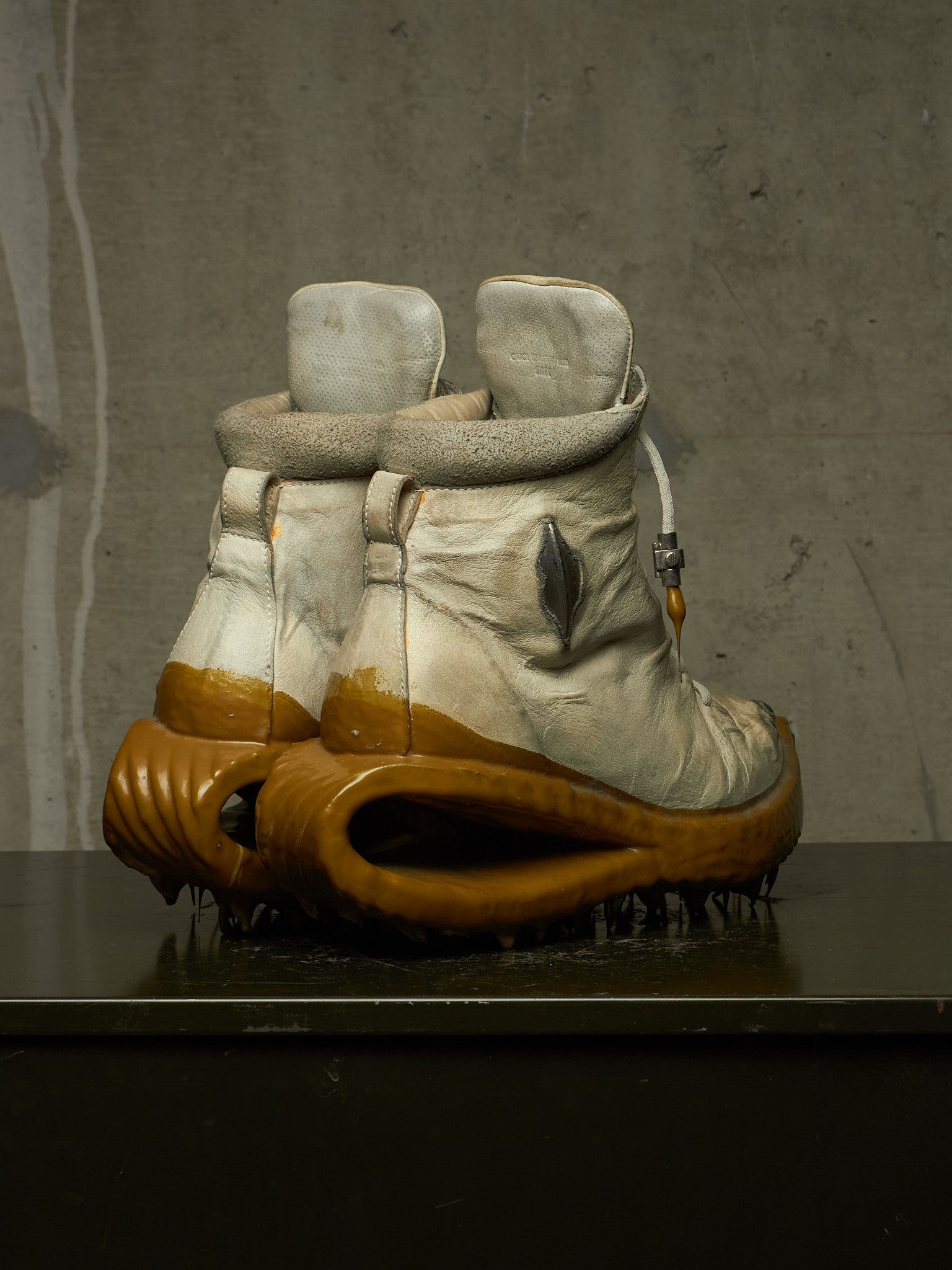 A pair of highly sculptural, distressed winter boots with off-white leather uppers, fuzzy gray trim, and thick, organic-shaped brown soles, displayed on a dark surface against a textured gray concrete wall.
