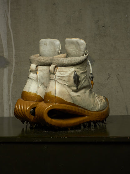 A pair of highly sculptural, distressed winter boots with off-white leather uppers, fuzzy gray trim, and thick, organic-shaped brown soles, displayed on a dark surface against a textured gray concrete wall.