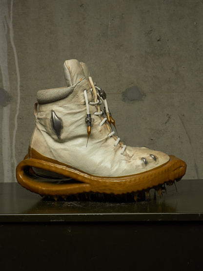 A single high-top boot with a distressed off-white leather upper, white laces, and a thick brown sculptural sole, displayed on a dark surface against a textured gray wall.
