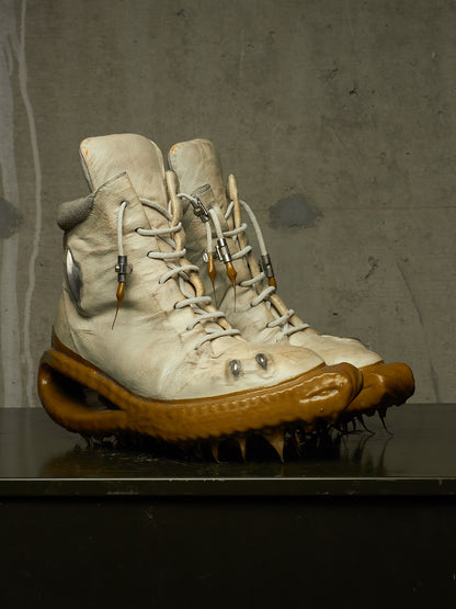 A pair of off-white high-top lace-up boots with thick brown spiked soles, displayed on a dark reflective surface against a textured gray wall.
