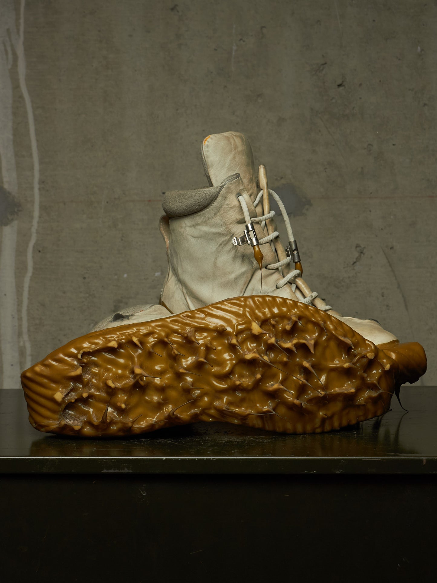 A sculptural art piece featuring a sculpted beige boot with white laces and a large, textured brown sole resting in the foreground against a dark gray concrete wall.