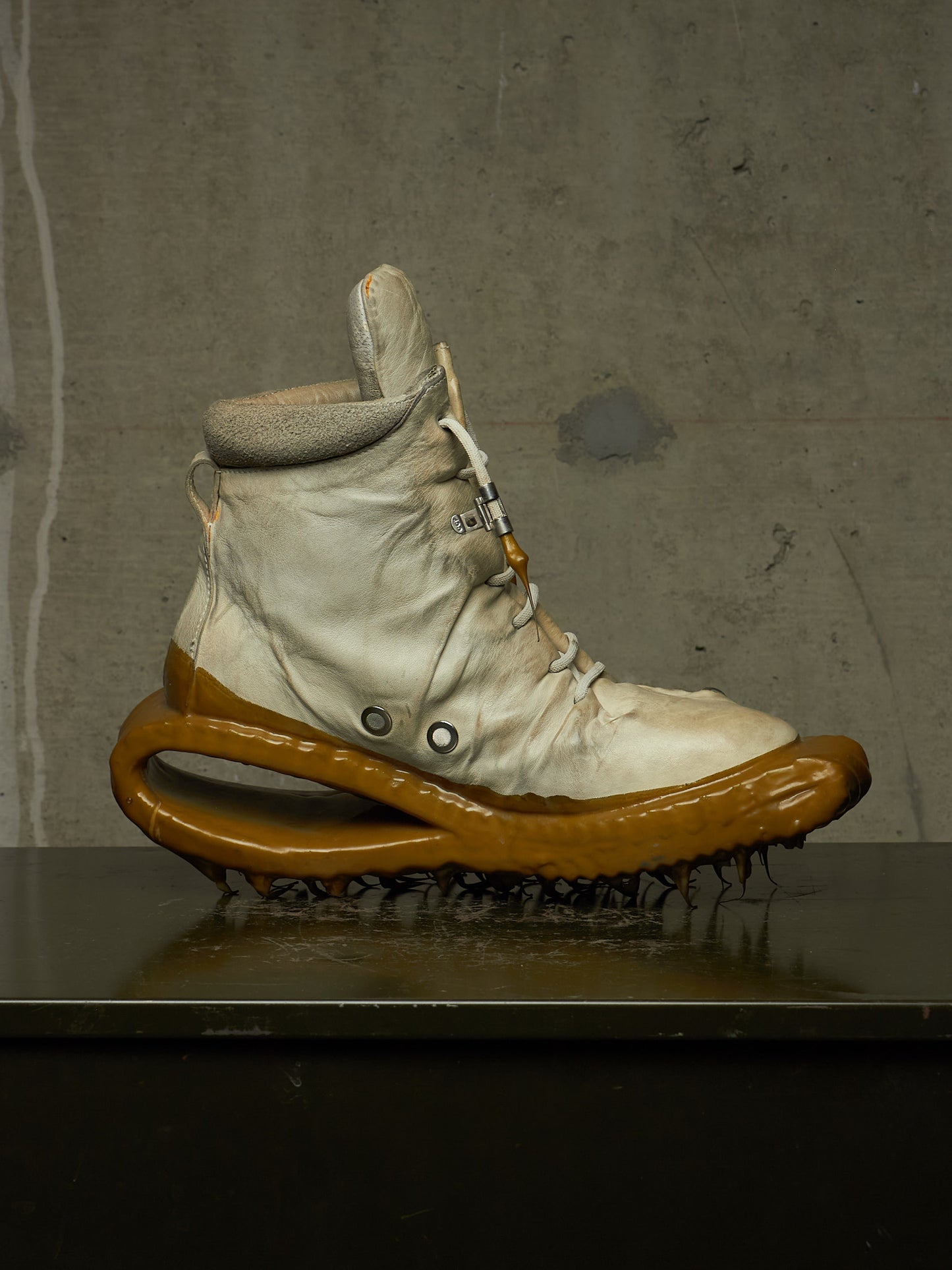 A single high-top sneaker with a cream-colored upper and a thick, sculptural glossy brown sole, displayed on a dark reflective surface against a textured gray concrete wall.