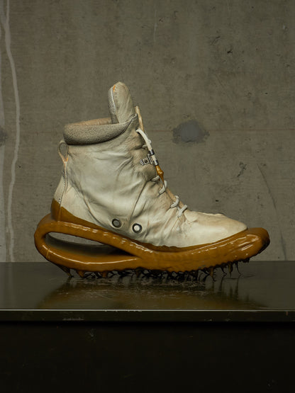 A single high-top sneaker with a cream-colored upper and a thick, sculptural glossy brown sole, displayed on a dark reflective surface against a textured gray concrete wall.