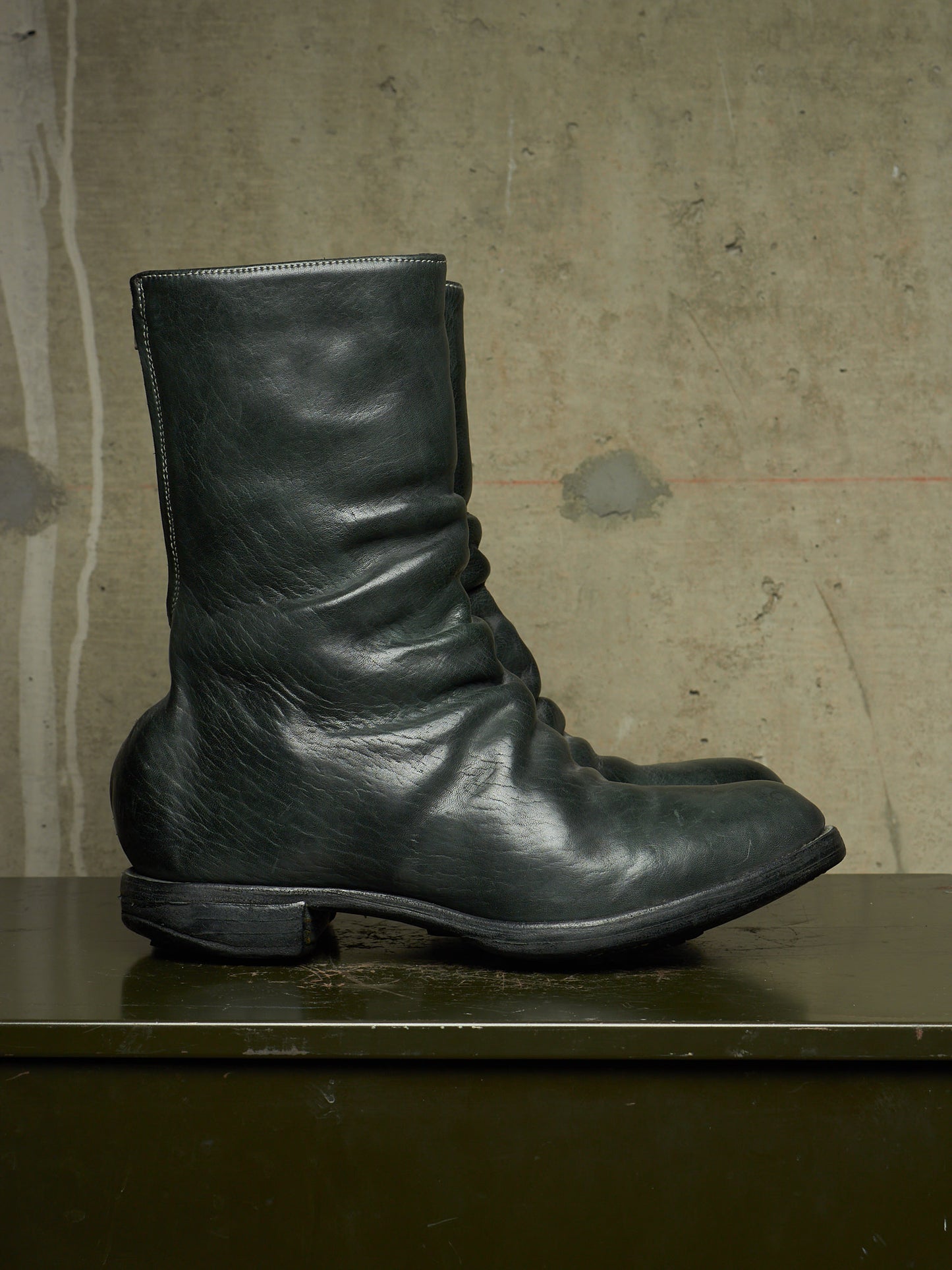 A single black leather ankle boot with a slouchy, gathered silhouette and low block heel, resting on a dark surface against a textured concrete wall.