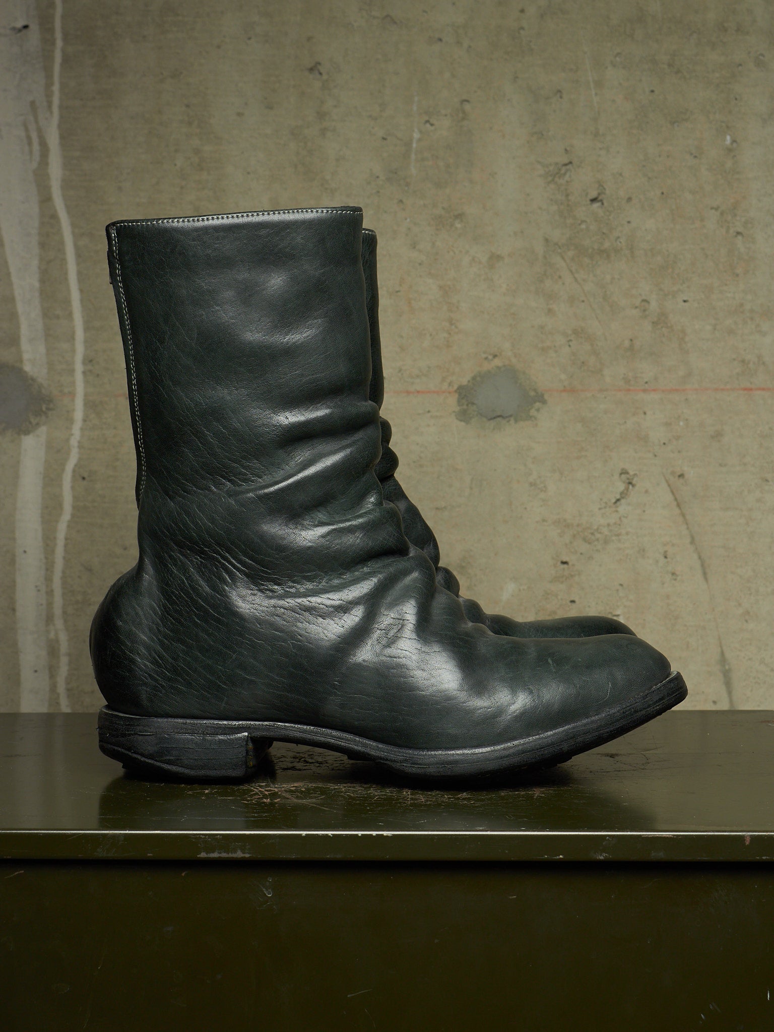 A single black leather ankle boot with a slouchy, gathered silhouette and low block heel, resting on a dark surface against a textured concrete wall.