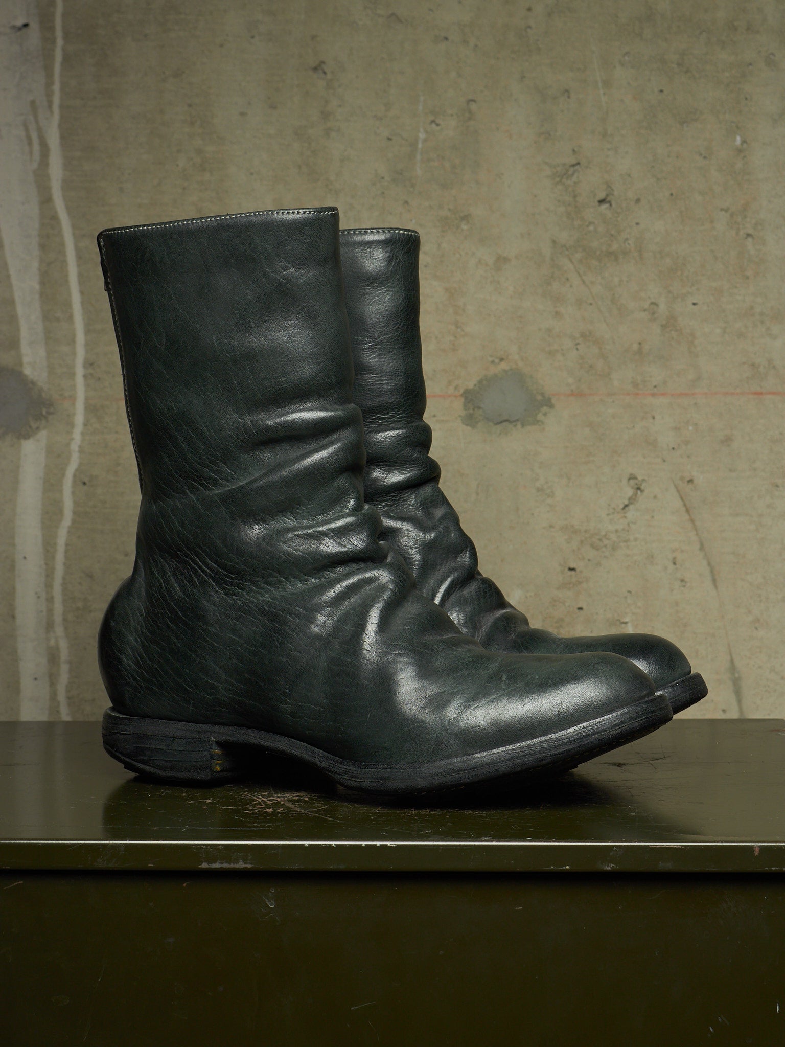 A pair of dark green, slouchy leather ankle boots resting on a dark green wooden surface against a textured concrete wall.