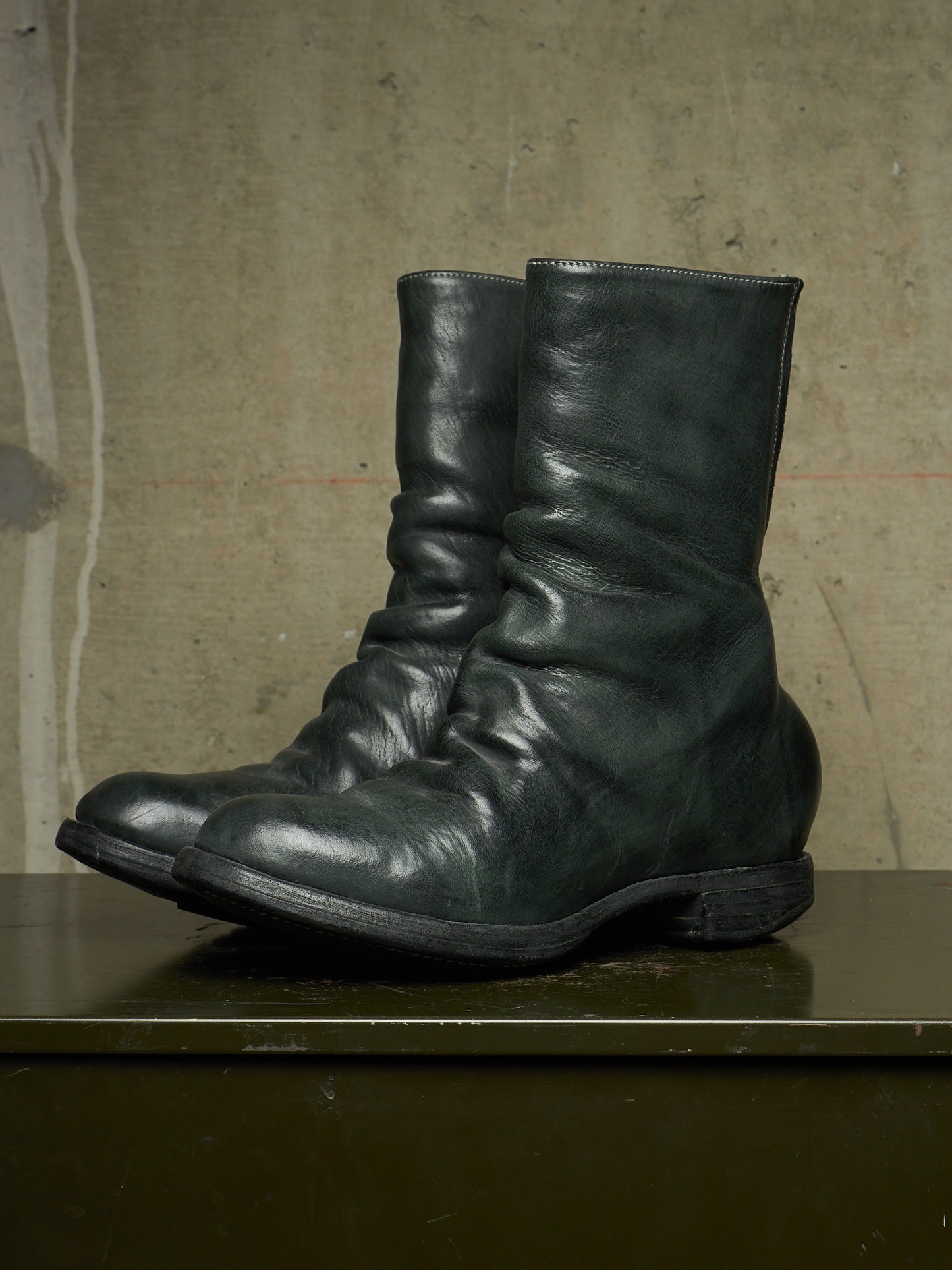 A pair of dark green leather mid-calf boots with a wrinkled, slouchy texture, resting on a dark green surface against a textured concrete wall.