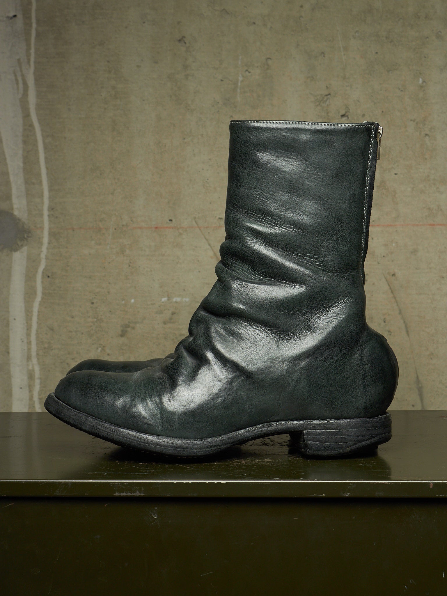 A single dark green leather mid-calf boot with a slouchy, gathered shaft and low block heel, resting on a dark green surface against a textured concrete wall.