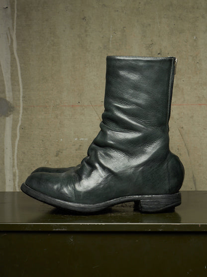 A single dark green leather mid-calf boot with a slouchy, gathered shaft and low block heel, resting on a dark green surface against a textured concrete wall.