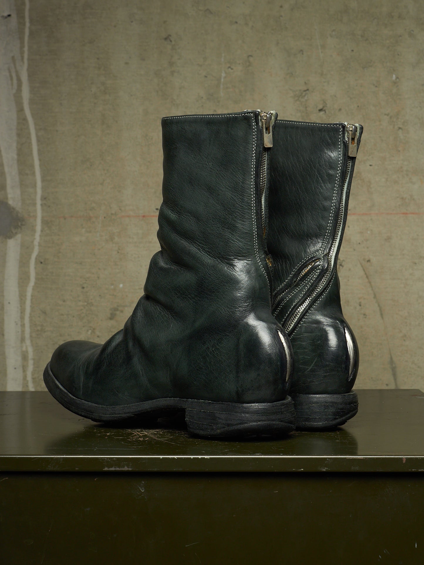 A pair of dark green leather ankle boots with side zippers, displayed on a dark surface against a textured concrete wall.