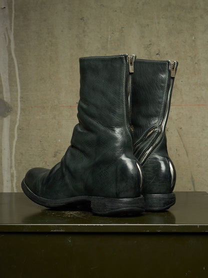 A pair of dark green leather ankle boots with side zippers, displayed on a dark surface against a textured concrete wall.