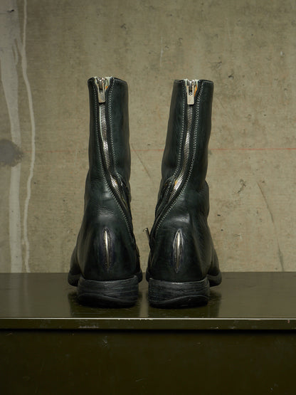 A pair of black leather ankle boots shown from the back, featuring a prominent silver zipper running up the back of each boot, set against a textured concrete wall.