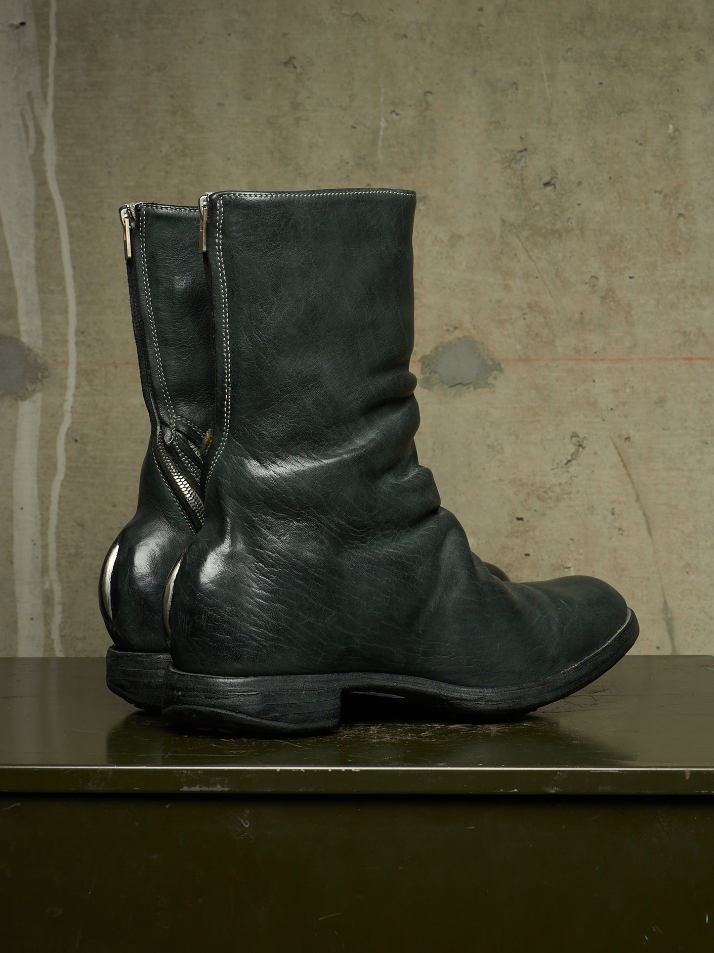 A pair of dark green leather mid-calf boots with side zippers, displayed on a dark green surface against a textured concrete wall.
