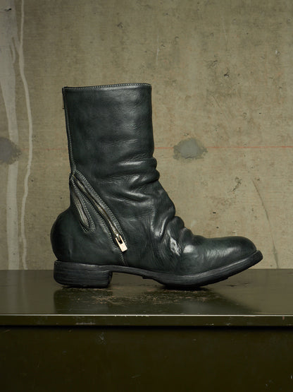 A single black leather ankle boot with a side zipper and a small silver metal pull tab, displayed in profile on a dark surface against a textured concrete wall.