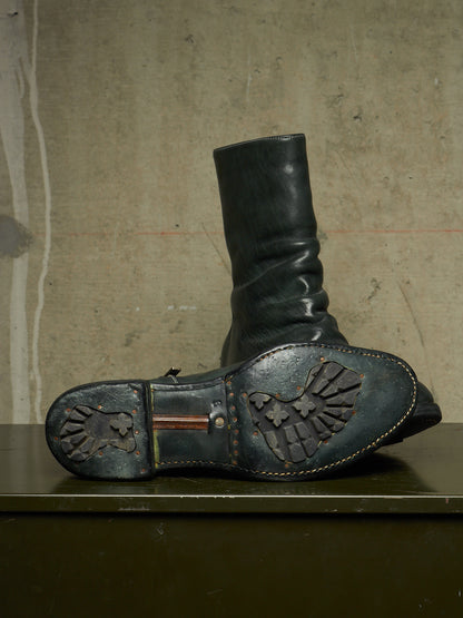 A single dark leather boot standing upright on a dark surface, with the sole turned up to show a heavily treaded, weathered rubber outsole against a textured concrete wall.