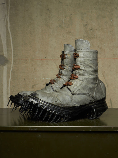 A pair of distressed gray combat boots with brown laces and spiked black soles, displayed on a dark surface against a textured beige wall.