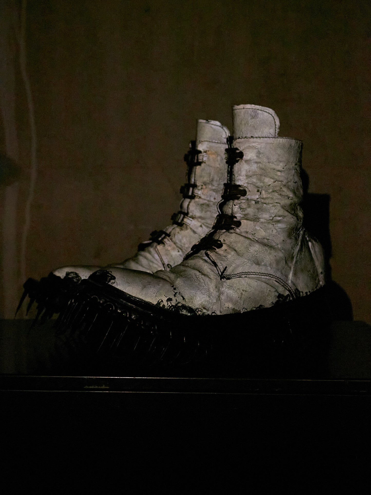 A pair of heavily distressed white lace-up combat boots with black spiked soles, photographed in a dark, moody setting against a brownish background.