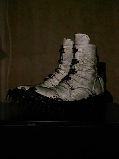 A pair of heavily distressed white lace-up combat boots with black spiked soles, photographed in a dark, moody setting against a brownish background.