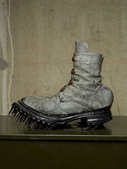A single distressed gray combat boot with a spiked black sole, displayed on a dark surface against a textured concrete wall.