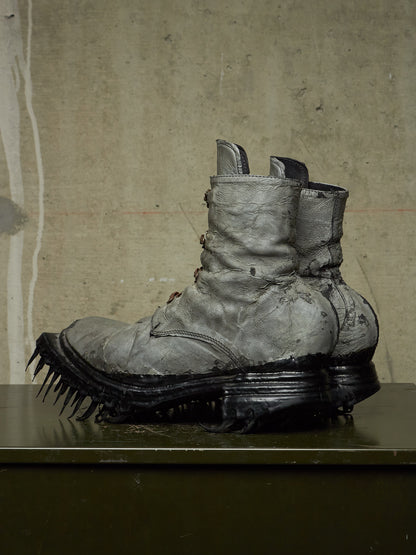 A pair of heavily distressed, oversized combat-style boots with thick black spiked soles, displayed on a dark surface against a textured concrete wall.