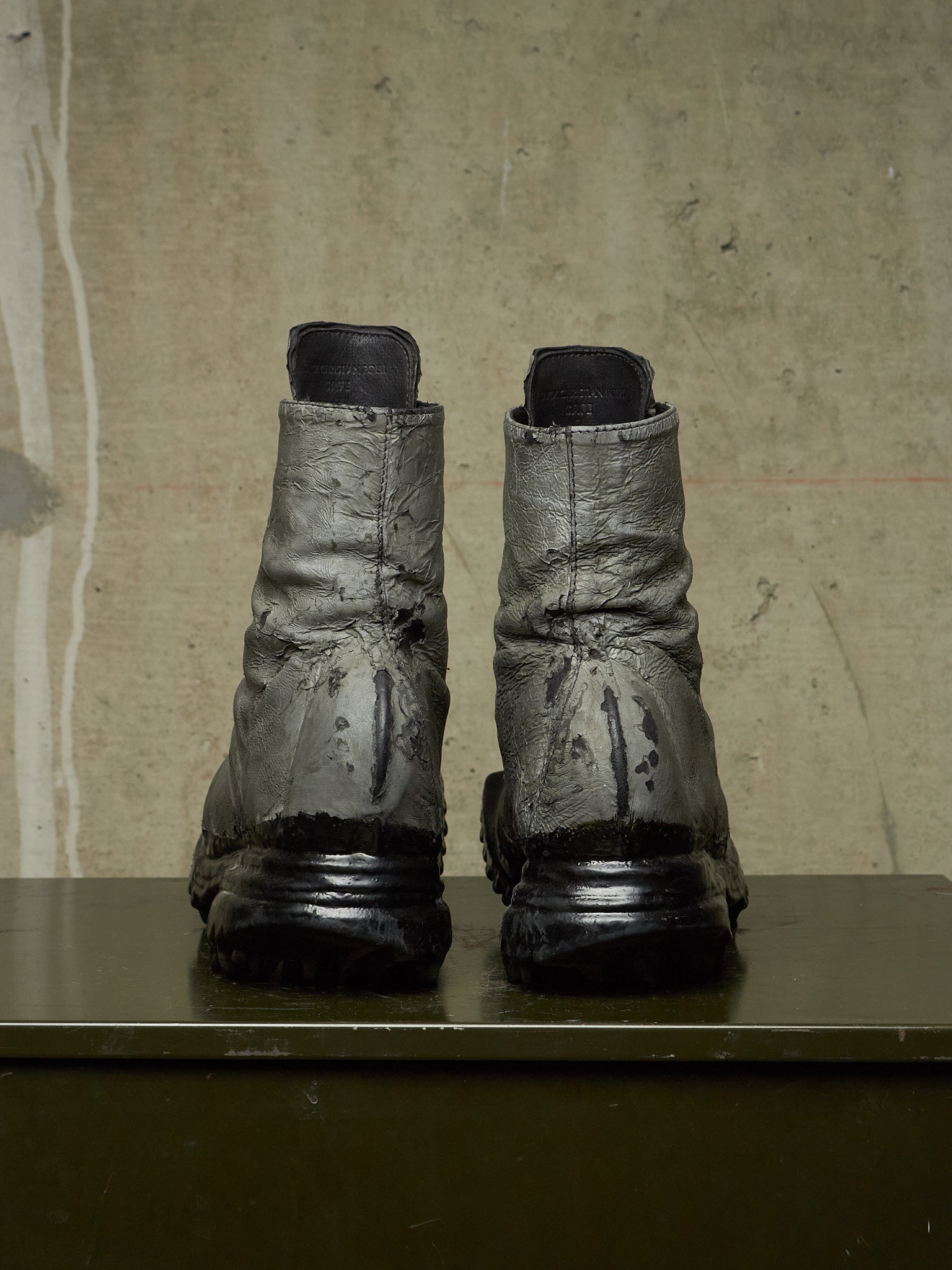 A pair of heavily distressed, dark metallic gray combat boots with black soles, standing side-by-side on a dark green surface against a textured beige concrete wall.
