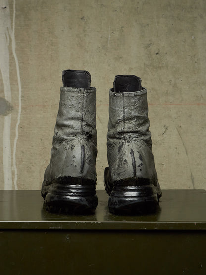 A pair of heavily distressed, dark metallic gray combat boots with black soles, standing side-by-side on a dark green surface against a textured beige concrete wall.