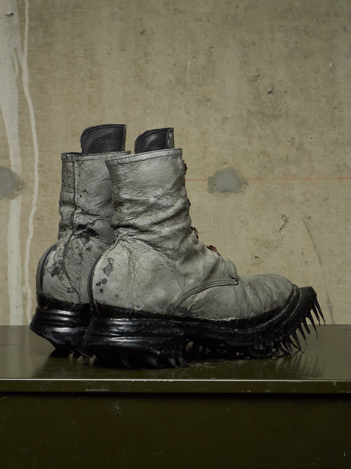 A pair of heavily distressed, oversized combat boots with thick black spiked soles, displayed on a dark surface against a textured concrete wall.