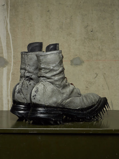 A pair of heavily distressed, oversized combat boots with thick black spiked soles, displayed on a dark surface against a textured concrete wall.