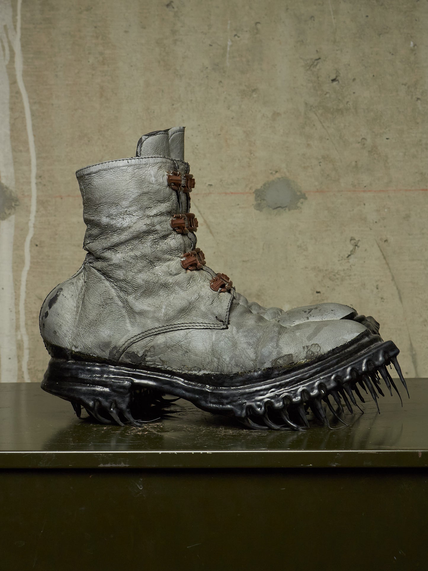 A single distressed gray combat-style boot with brown laces and a black spiked sole, displayed on a dark surface against a textured concrete wall.