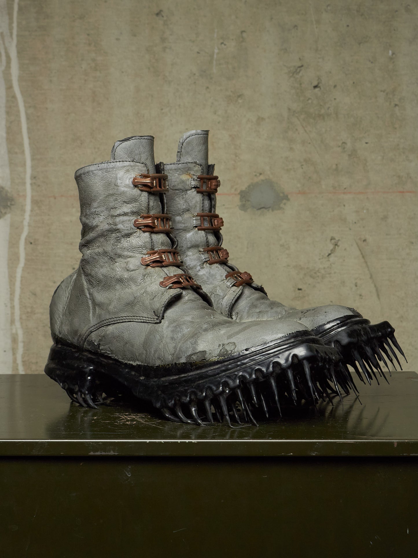 A pair of distressed gray combat boots with brown laces and spiked black soles, displayed on a dark reflective surface against a textured concrete wall.