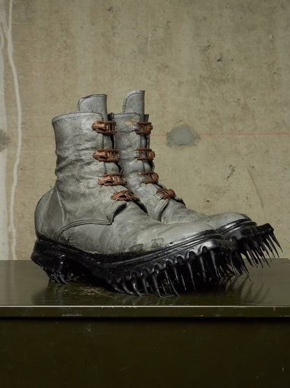 A pair of distressed gray combat boots with brown laces and spiked black soles, displayed on a dark reflective surface against a textured concrete wall.