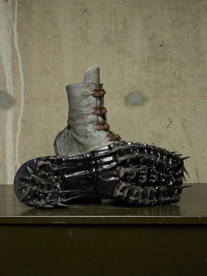 A unique sculptural combat boot featuring a textured gray leather upper with brown laces and a black spiked, treaded sole, displayed on a dark green table against a concrete wall.