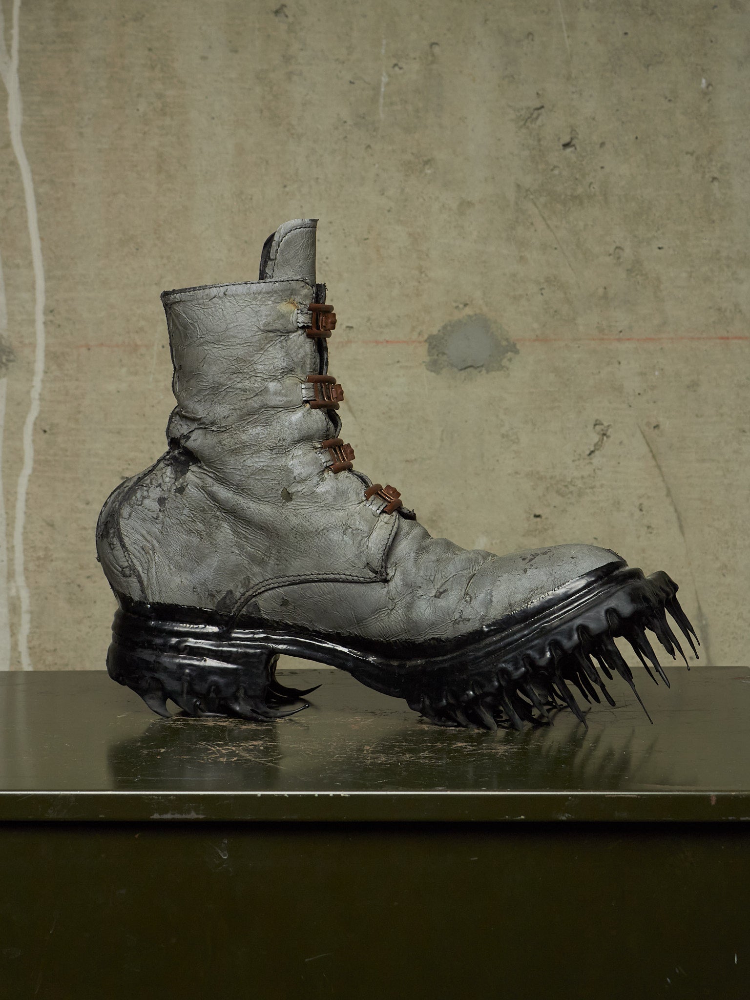 A single distressed gray leather lace-up boot with a chunky black spiked sole, displayed on a dark reflective surface against a textured concrete wall.