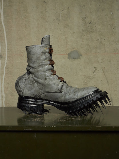 A single distressed gray leather lace-up boot with a chunky black spiked sole, displayed on a dark reflective surface against a textured concrete wall.