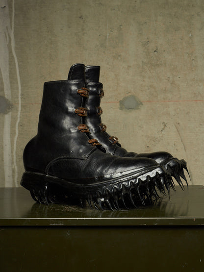 A pair of black leather combat boots with spiked soles and brown laces, displayed on a dark reflective surface against a textured concrete wall.