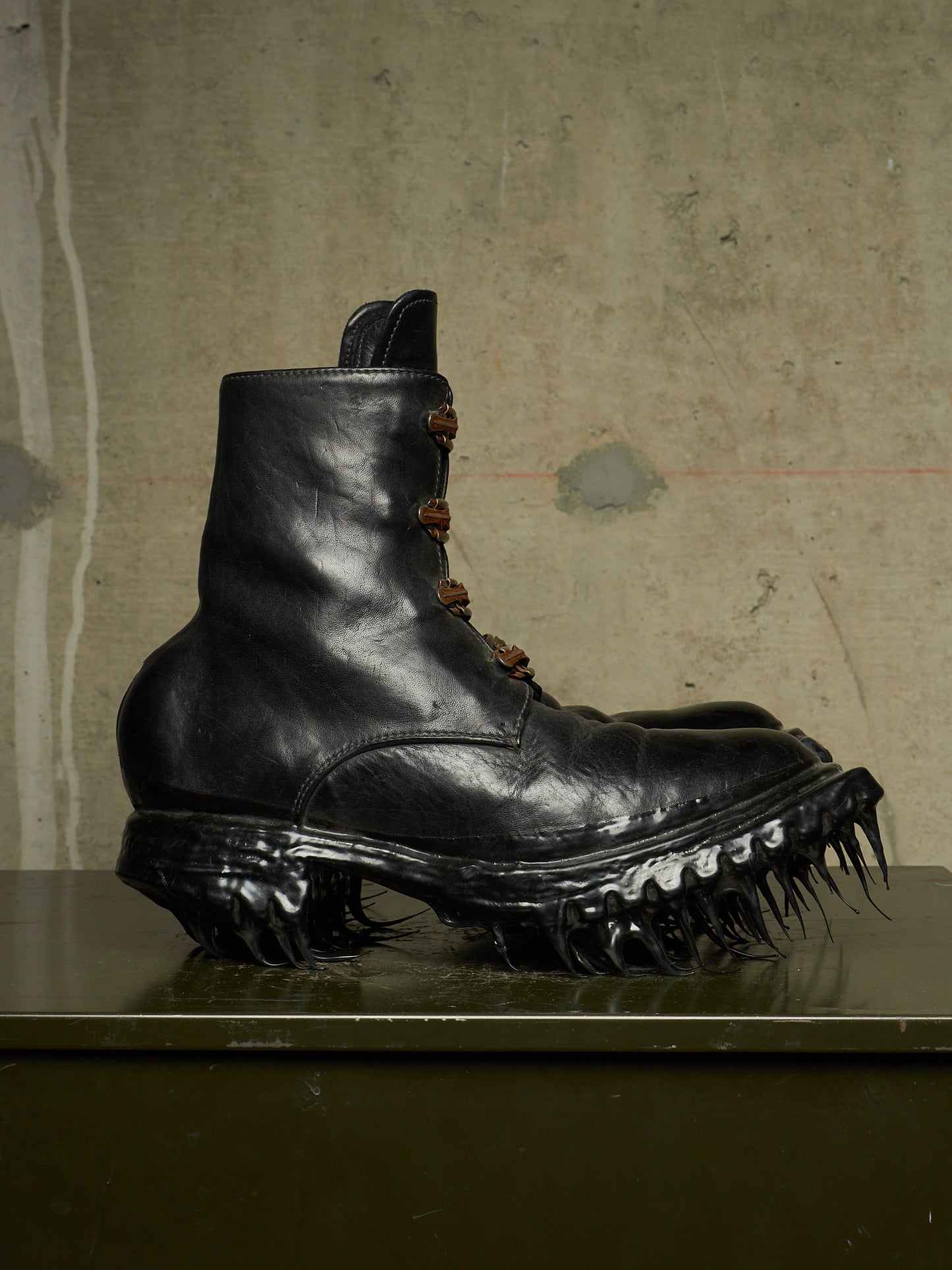 A black leather combat boot with a spiked, claw-like sole, displayed on a dark surface against a textured concrete wall.