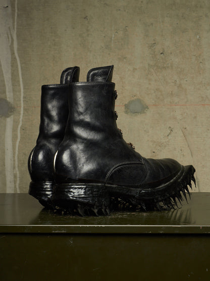 A pair of black leather ankle boots with spiked, treaded soles, displayed on a dark surface against a textured concrete wall.