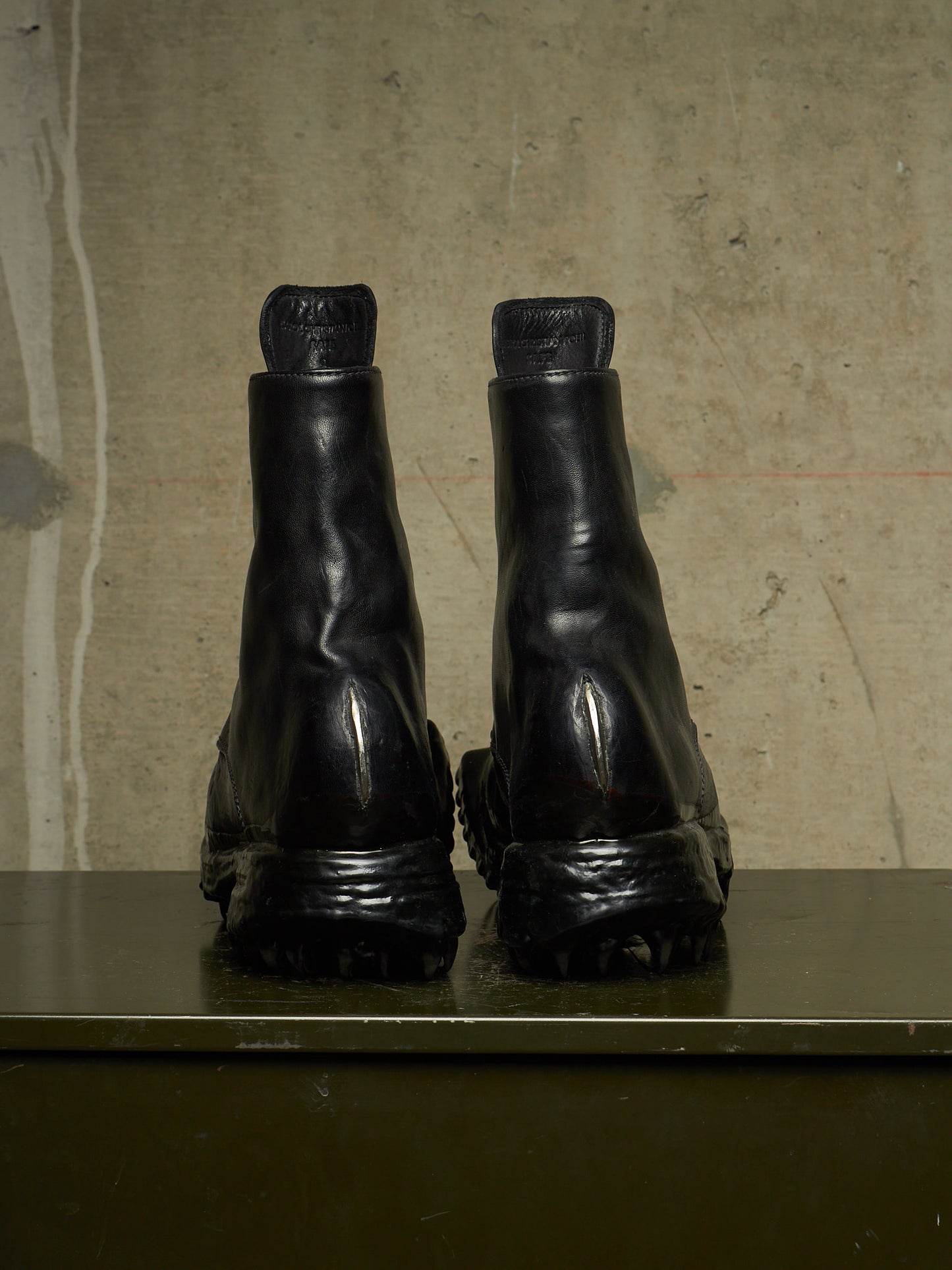 A pair of black leather Chelsea boots shown from the back, resting on a dark green surface against a textured beige concrete wall.