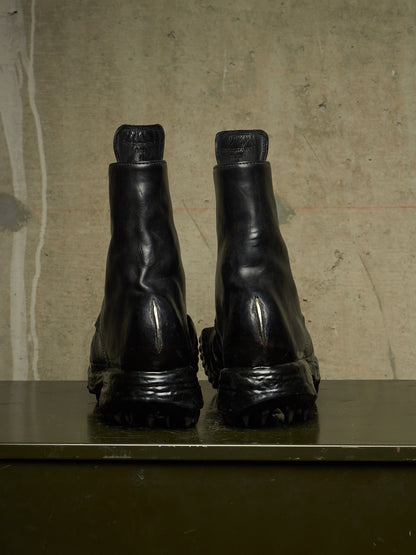 A pair of black leather Chelsea boots shown from the back, resting on a dark green surface against a textured beige concrete wall.