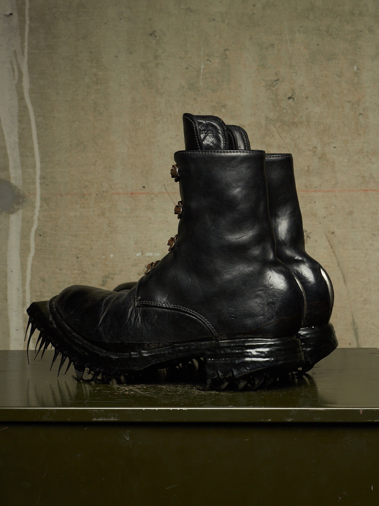 A pair of black leather combat boots with spiked lug soles, displayed on a dark reflective surface against a textured concrete wall.