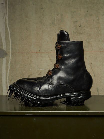 A black leather ankle boot with a heavily spiked lug sole, displayed on a dark surface against a textured concrete wall.